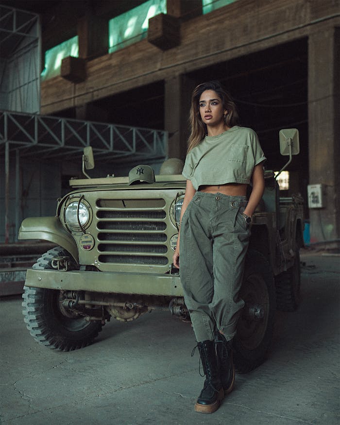 Stylish woman in khaki outfit poses confidently against a vintage jeep in an industrial warehouse.