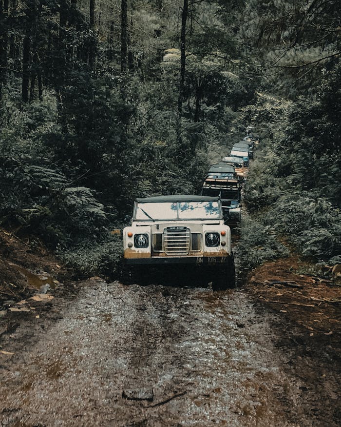 A convoy of vehicles navigates a muddy road through the lush forest of West Java, Indonesia.